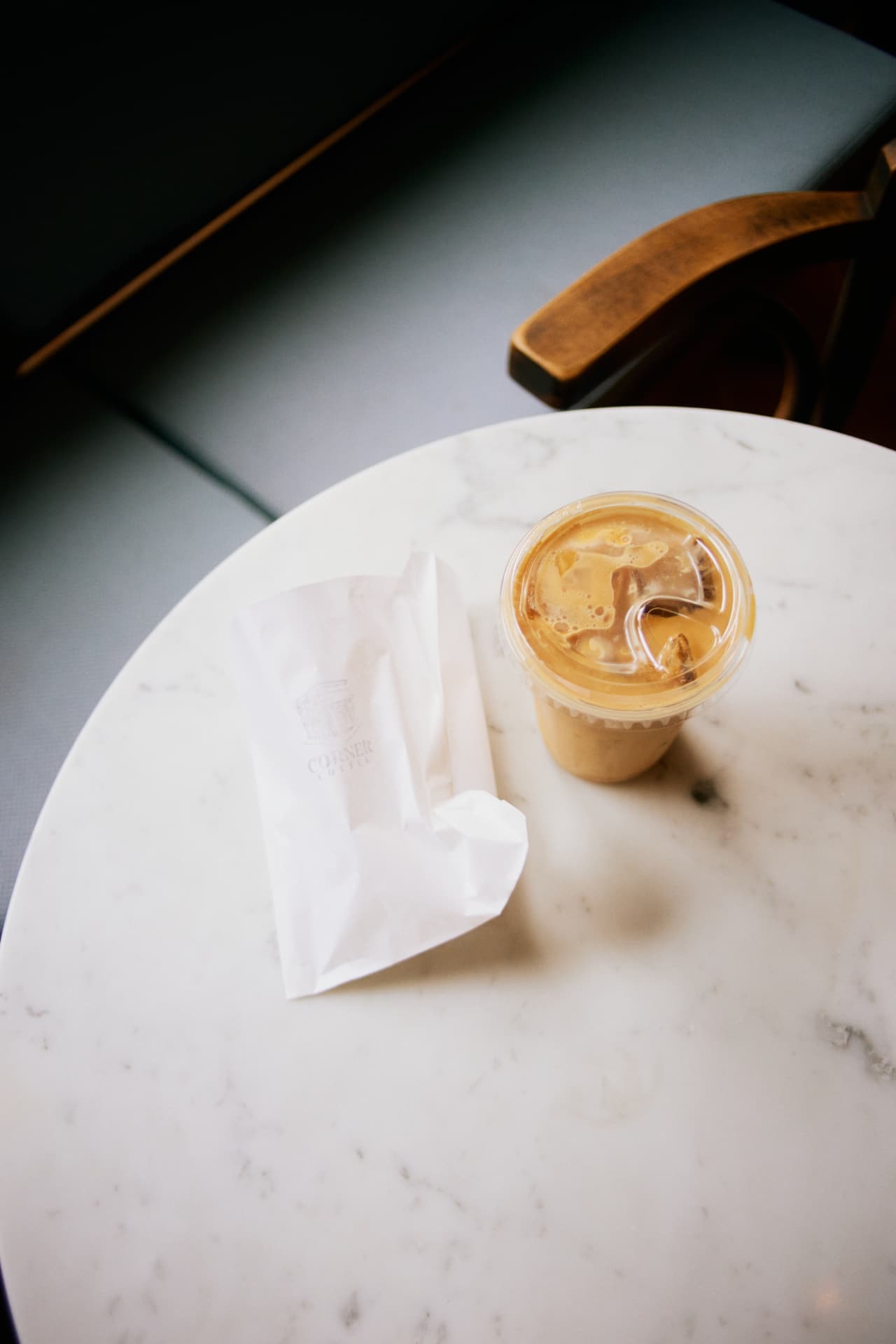 Iced coffee in a clear cup beside a Corner Coffee paper bag on a marble table, a teal banquette behind