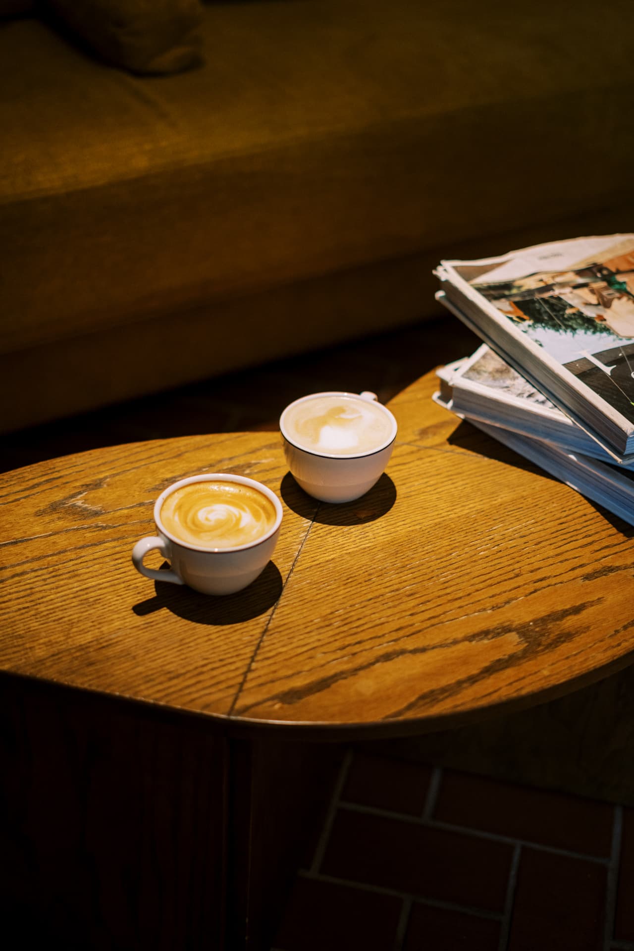 Two lattes with latte art on a curved wooden table beside a stack of magazines