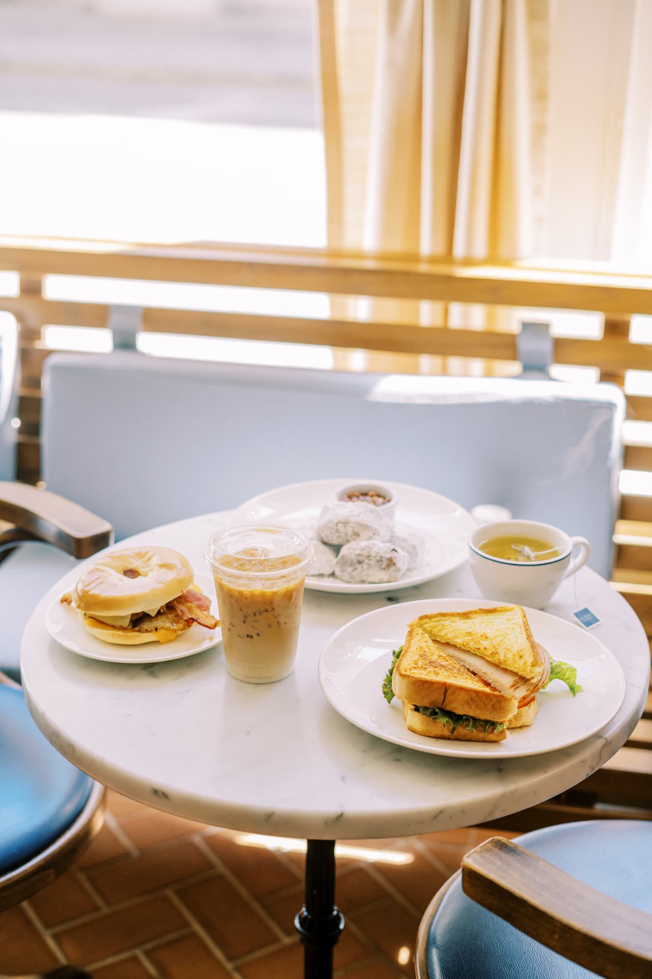 Marble bistro table with a bagel sandwich, a toasted deli sandwich, powdered beignets, an iced coffee and a cup of tea