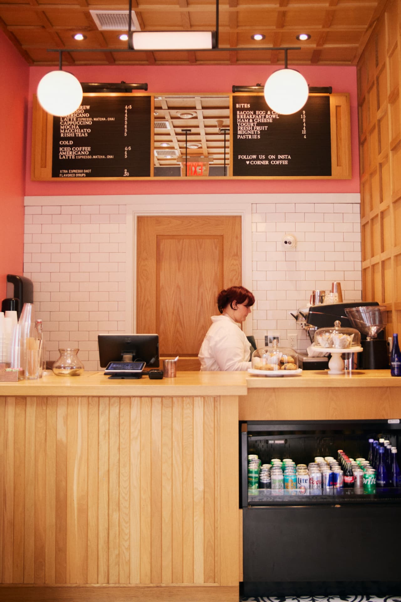 Barista at the service counter with pastries under glass cloches and a stocked drink fridge