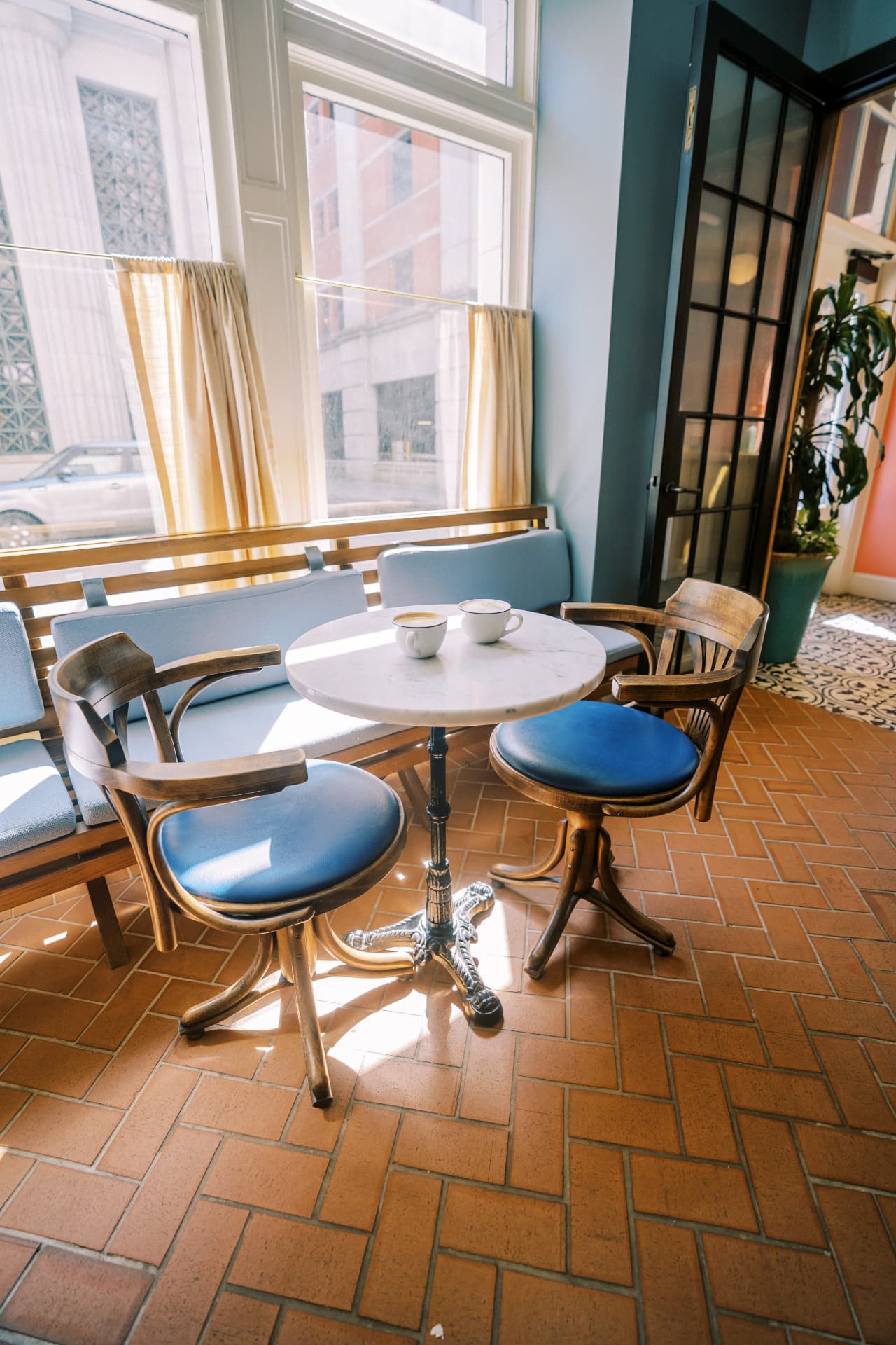Sunlit seating corner with a marble table holding two coffees, blue chairs and a bench under tall windows
