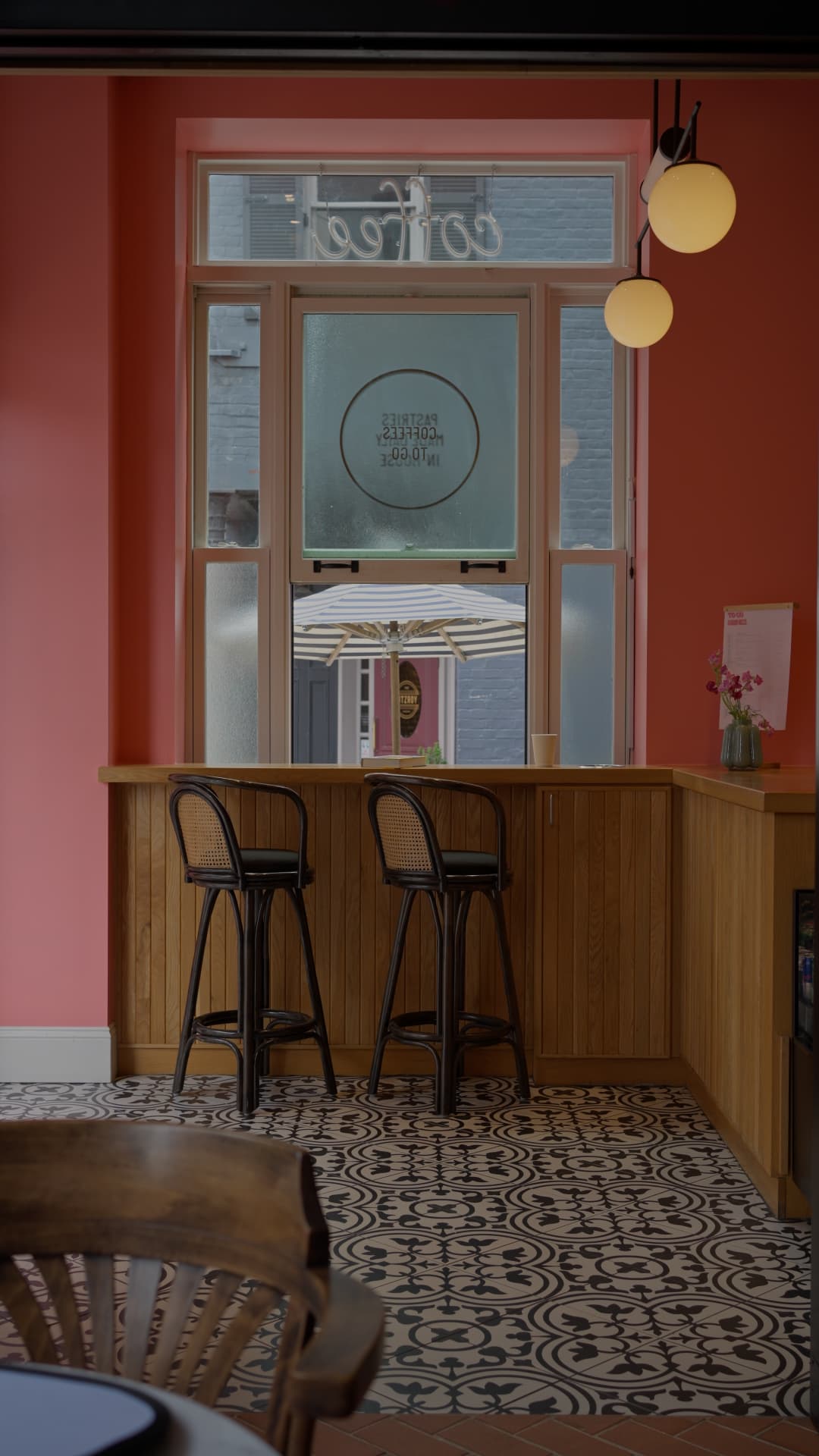 Cane barstools at the window counter with a takeaway cup, coral wall and patterned tile floor